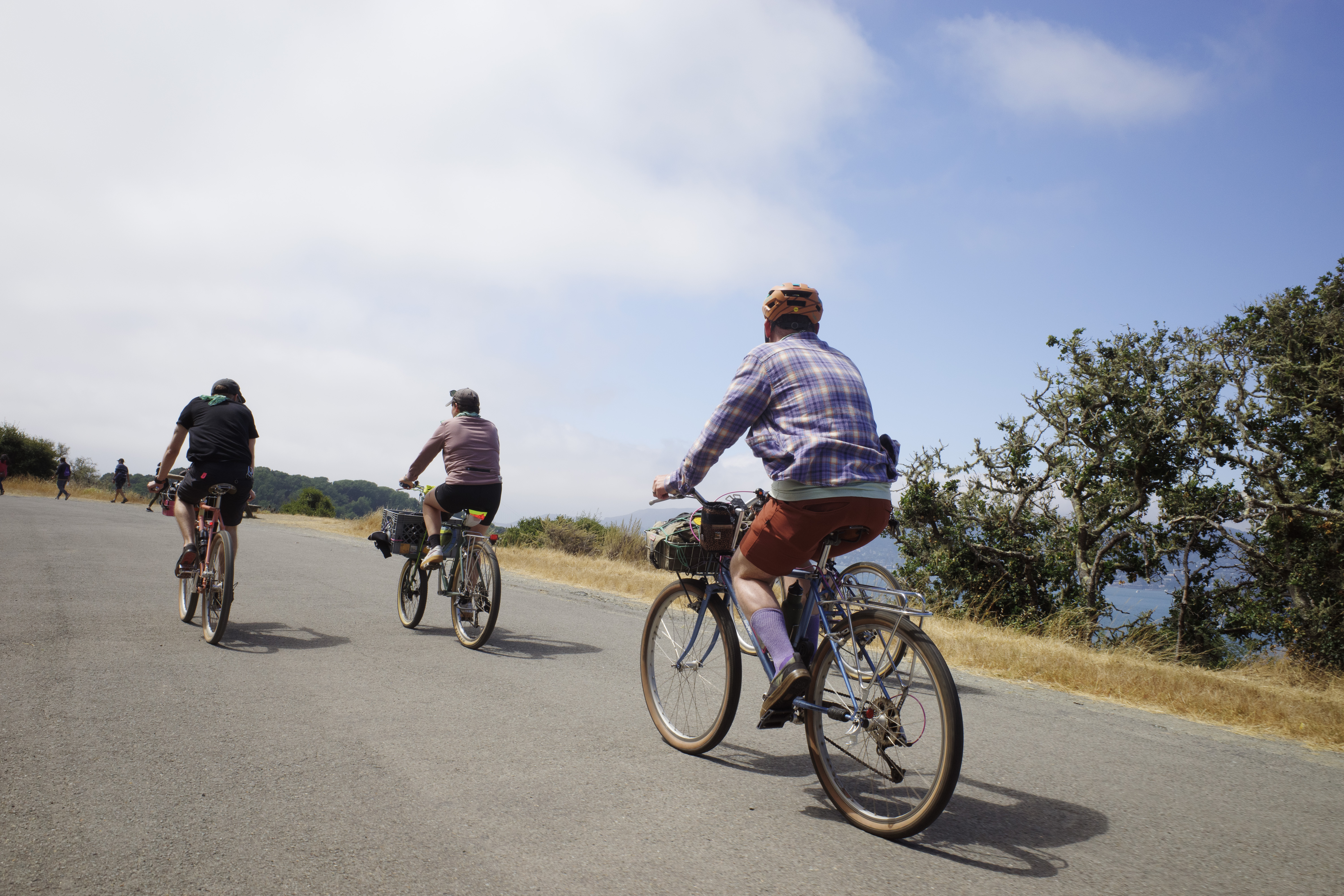 A cyclist wearing flannel biking up a paved hill with two other cyclists in front of him. The background has blue skies with large puffy white clouds.