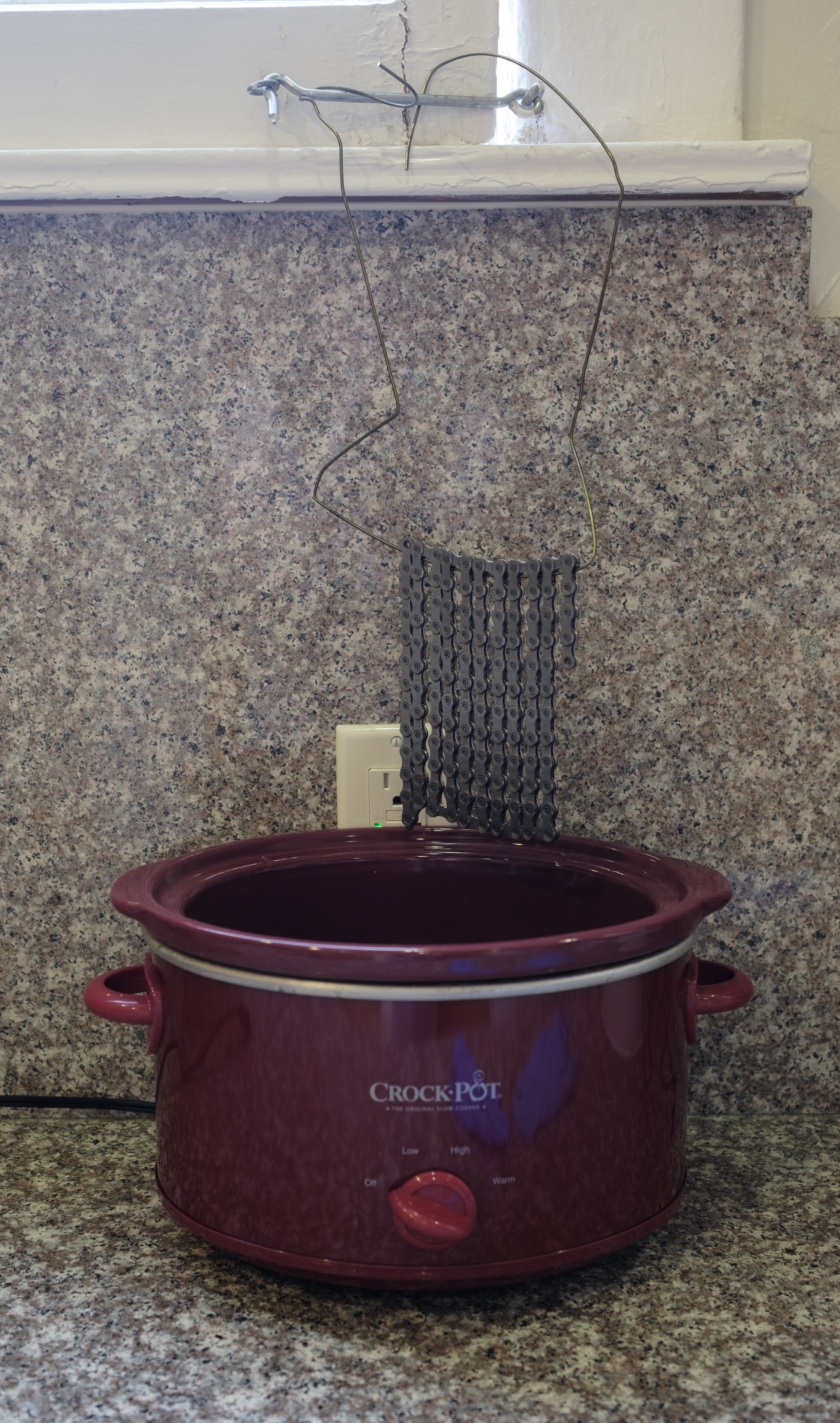 A chain suspended over a slow cooker via a clothes hanger hooked on a window latch