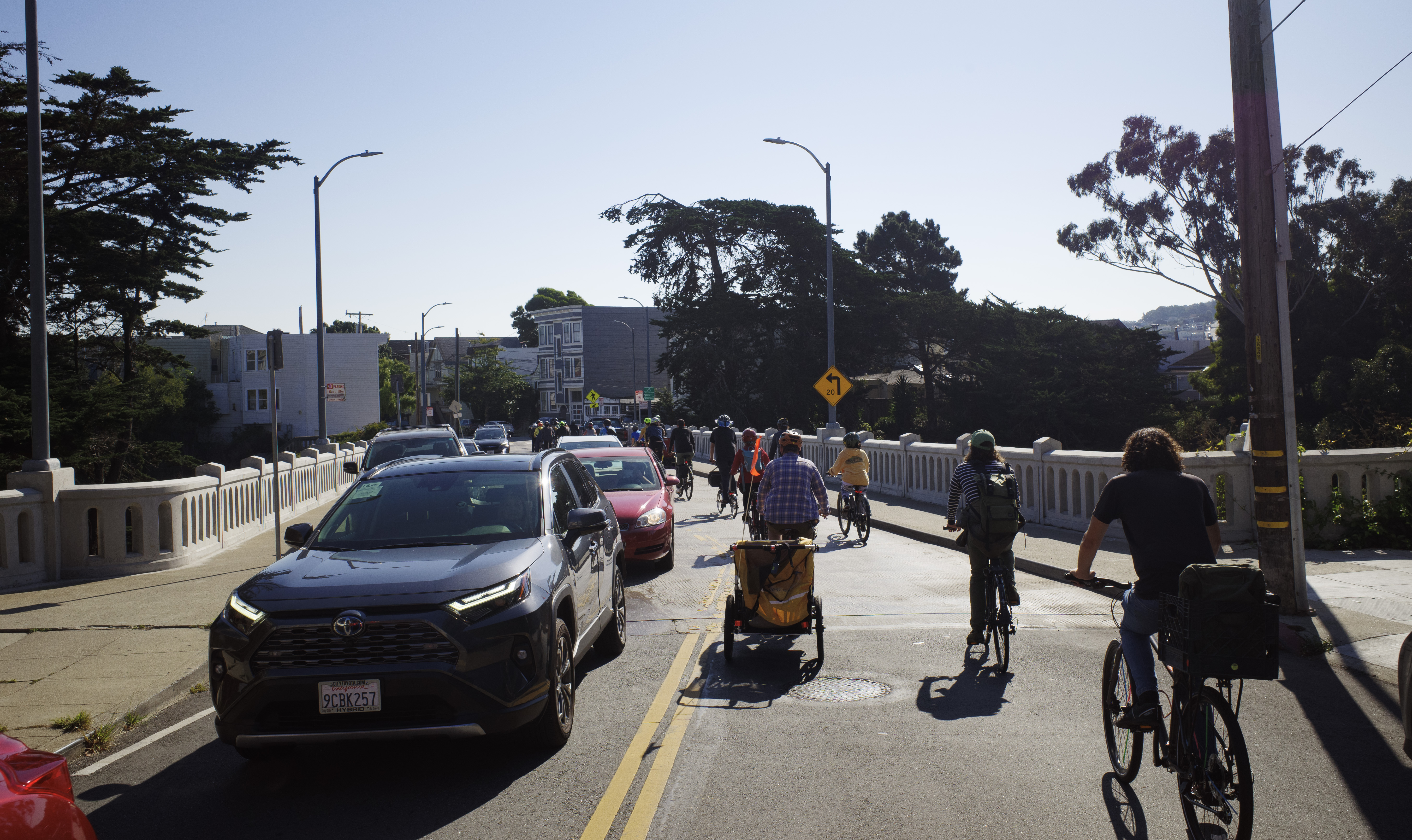A group of cyclists taking all the space on the right lane over a bridge, with a line of cars in the left lane