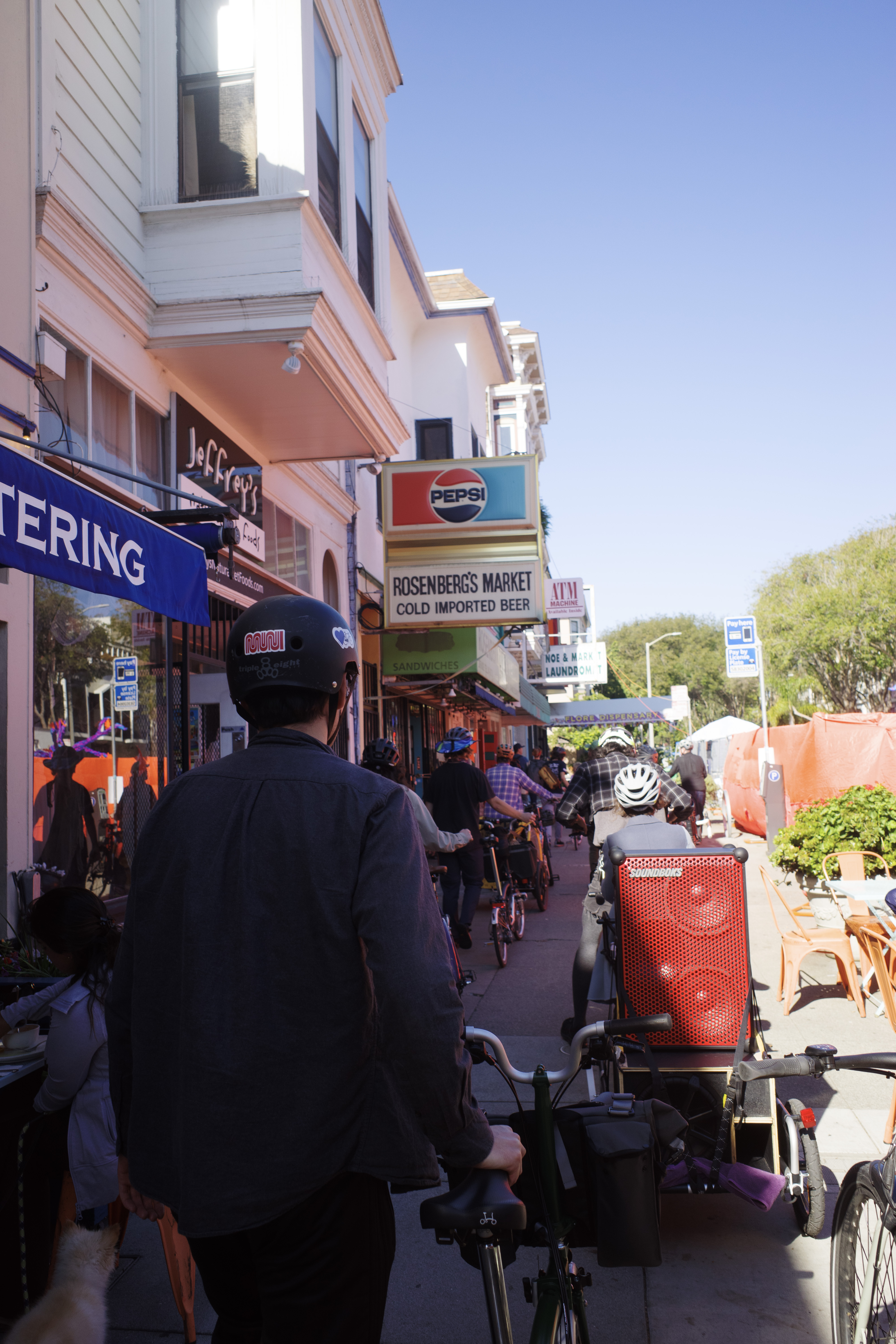 A group of cyclists walking their bikes on the sidewalk. The person in front is wearing a helmet with a MUNI worm sticker.