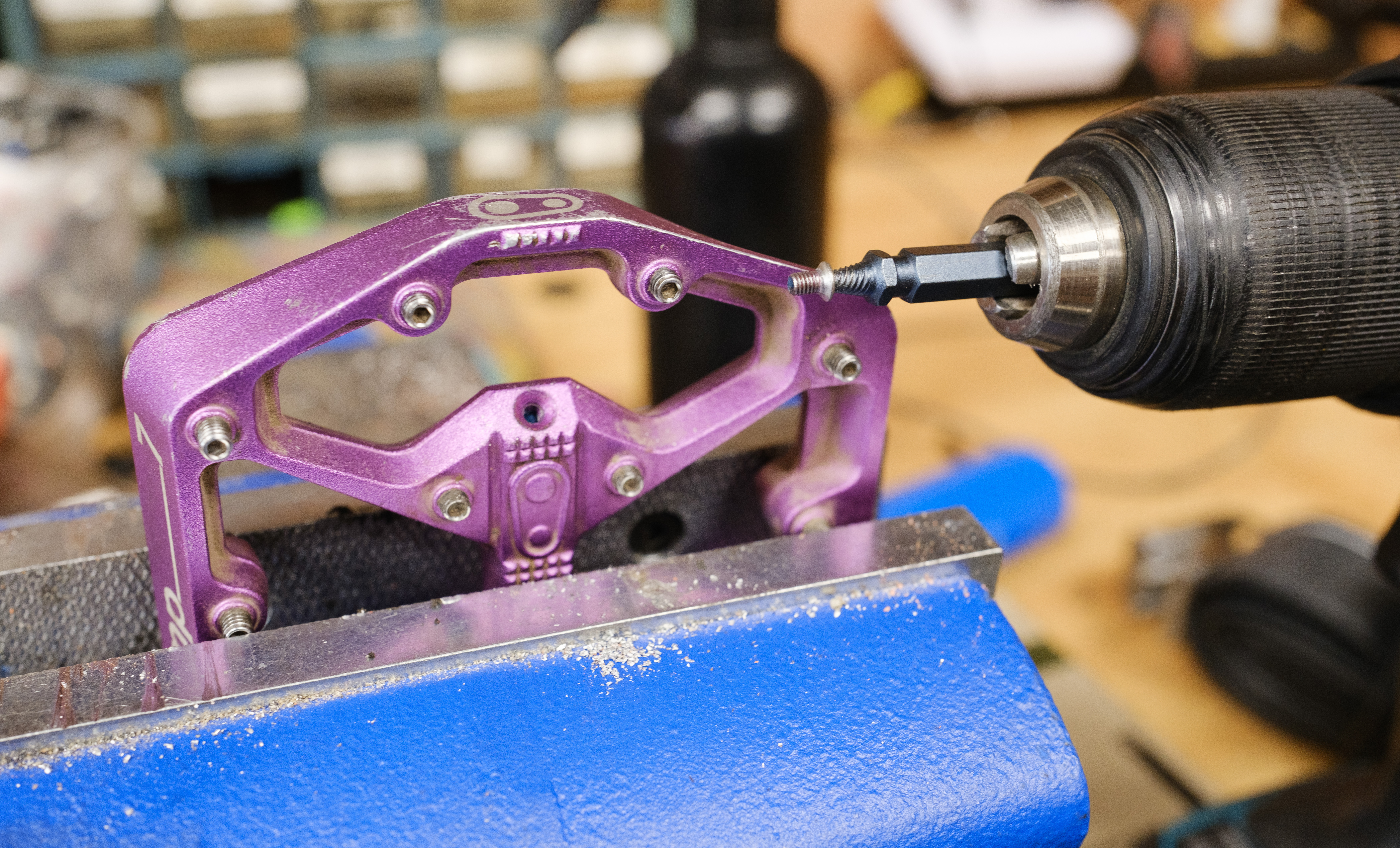 A Crankbrothers Stamp 7 left pedal in a bench vise beside a drill with a bolt extractor and grease port screw hanging on the bolt extractor