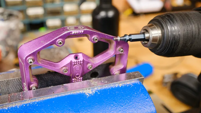 A Crankbrothers Stamp 7 left pedal in a bench vise beside a drill with a bolt extractor and grease port screw hanging on the bolt extractor