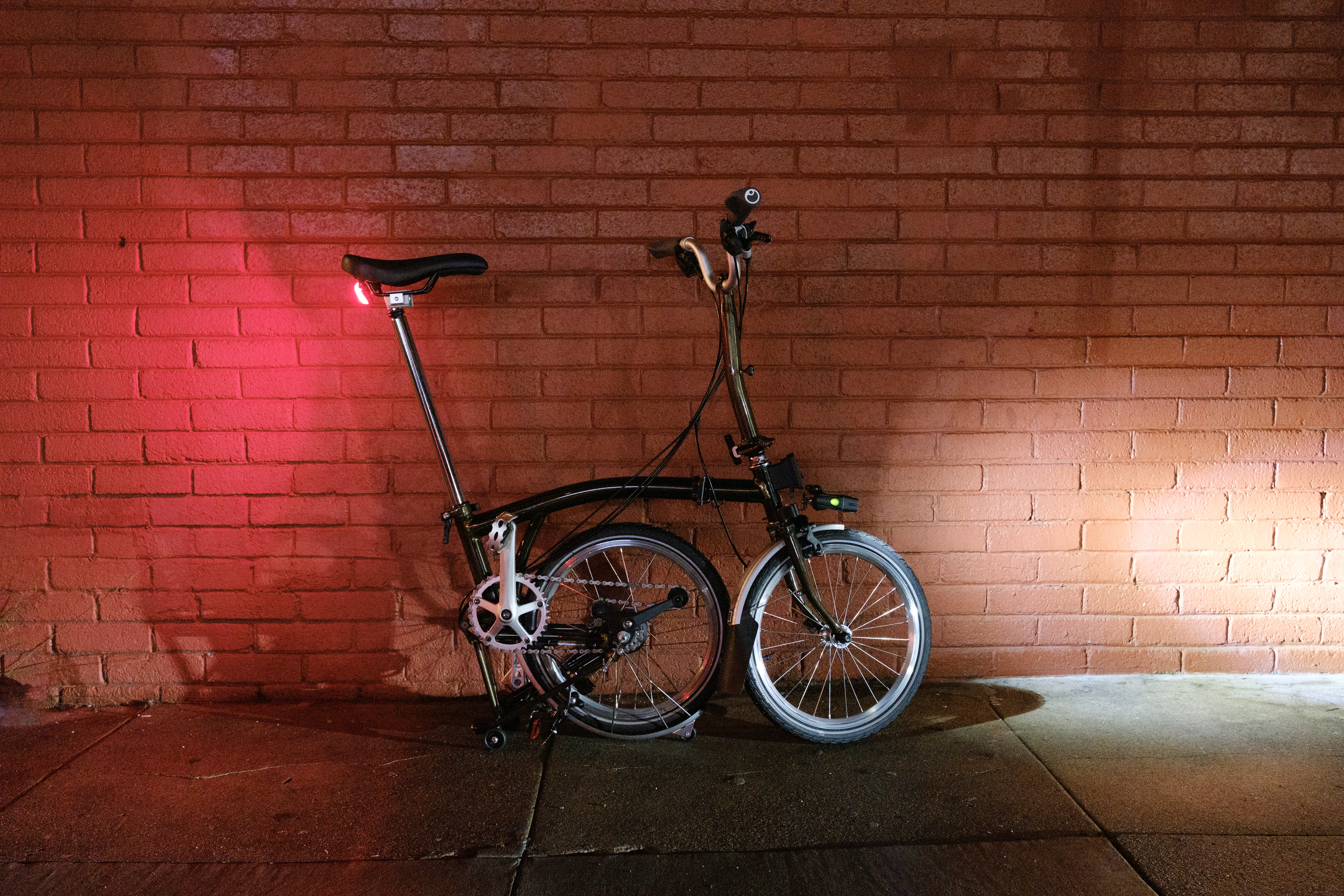 A semi-folded Brompton in the parked position in front of a brick wall at night.