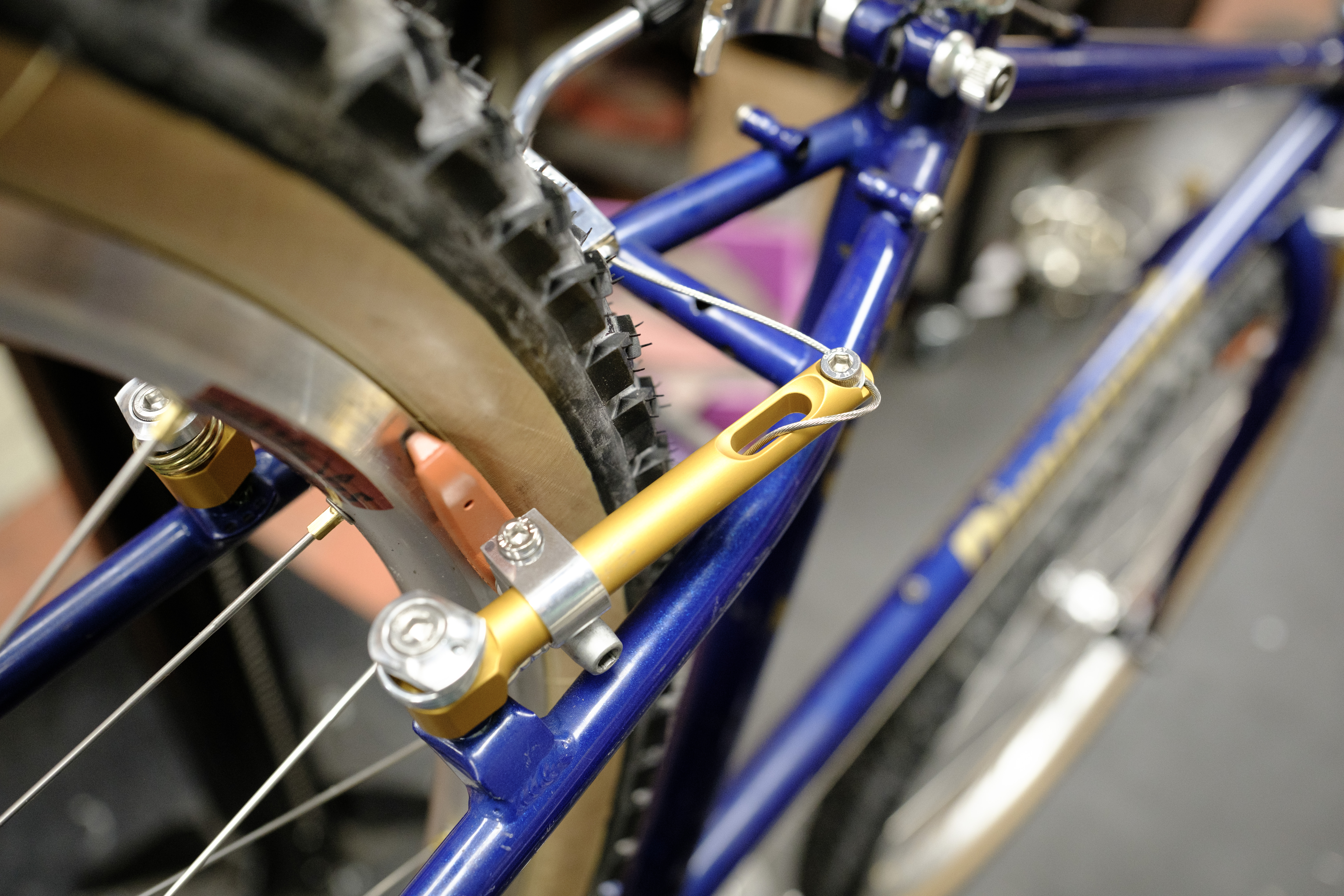 A close-up of a gold brake with the excess cable routed through its slot. It's mounted on a blue bike in a bike stand with a tanwall tire with black knobs.
