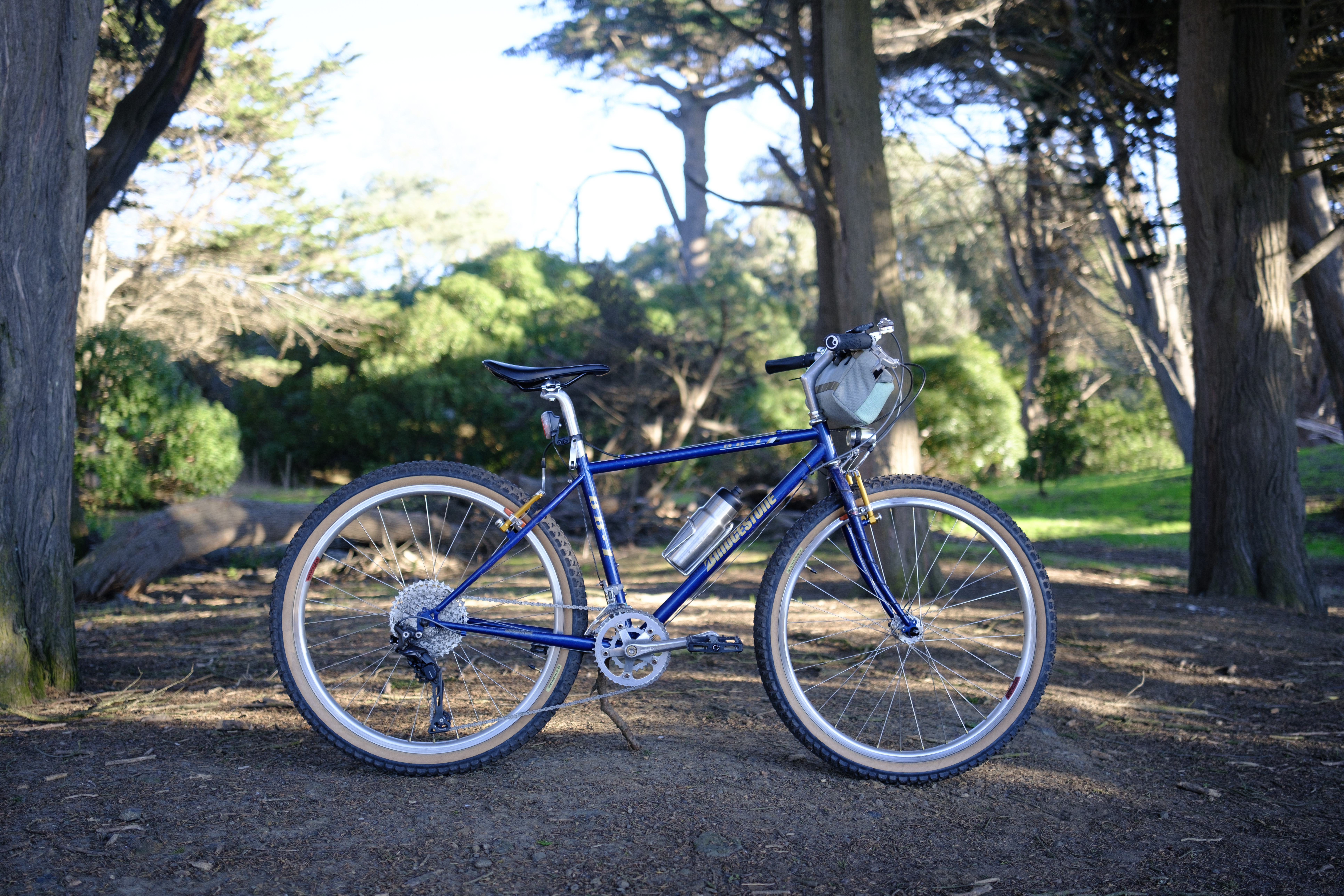 A blue bike with gold accents propped up with a stick on a dirt trail in a forest.