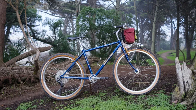 A blue and gold bicycle propped up with a stick in a misty forest. The bike has a dark tanwall rear tire and a medium tanwall front tire. There are silver cranks and a silver front rack with a burgundy bag.