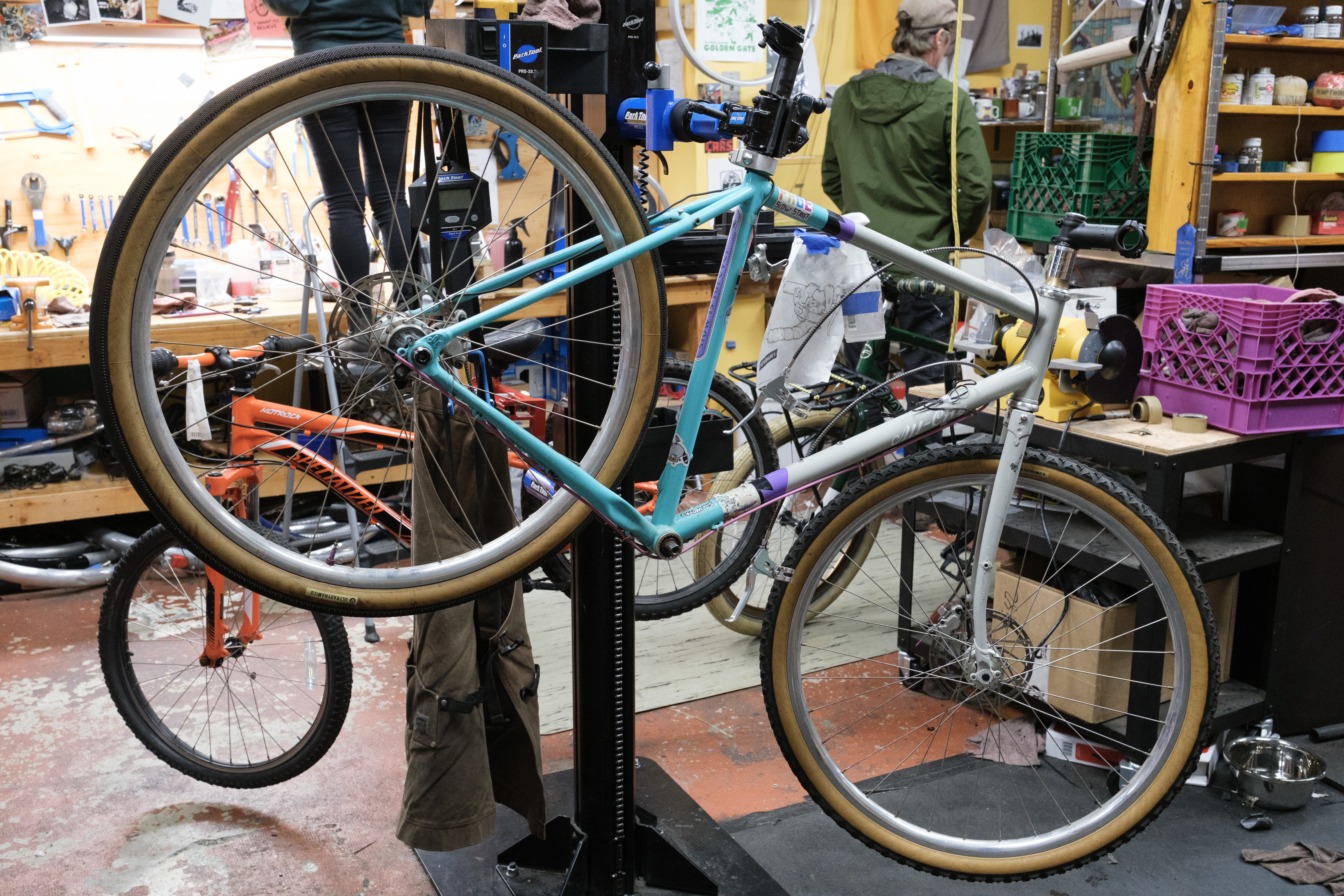 A grey/turquoise bicycle in a bike stand with only its wheels, bottom bracket, seatpost, and a stem.