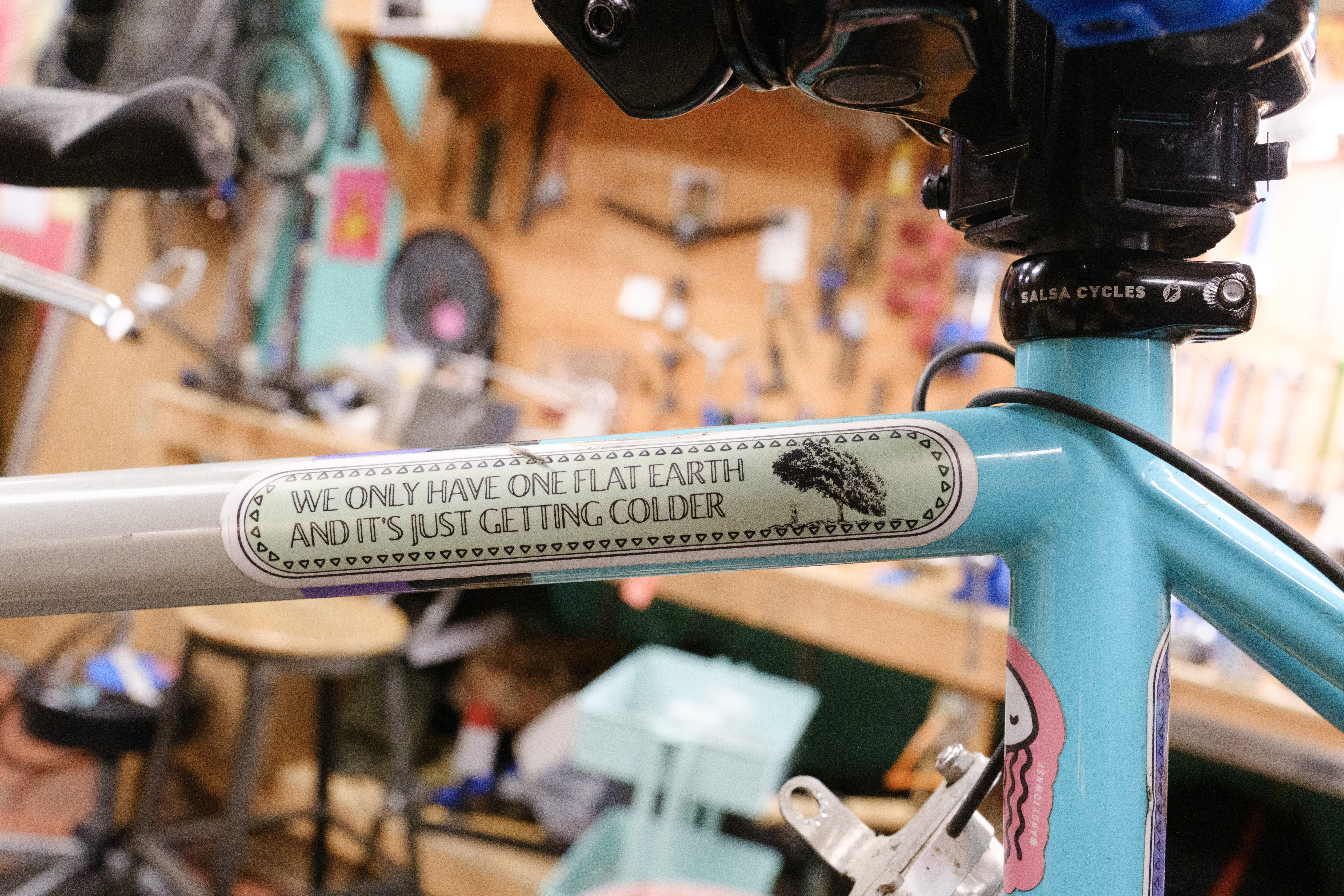 The top tube/seat tube junction of a bicycle in the stand with a Salsa Cycles seat collar from the previous picture and a sticker that reads 'we only have one flat earth and its just getting colder'.