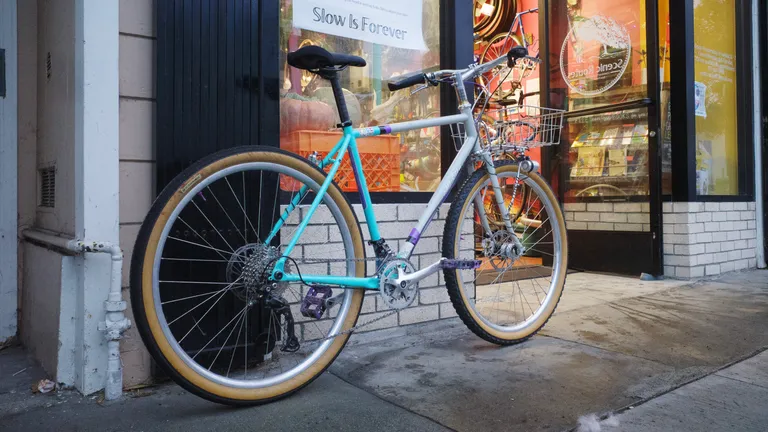 A bicycle with a grey/turquoise frame, black and tan tires, and a mix of black and silver components. It's leaning outside on the wall of Scenic Routes Community Bicycle Center in the evening.