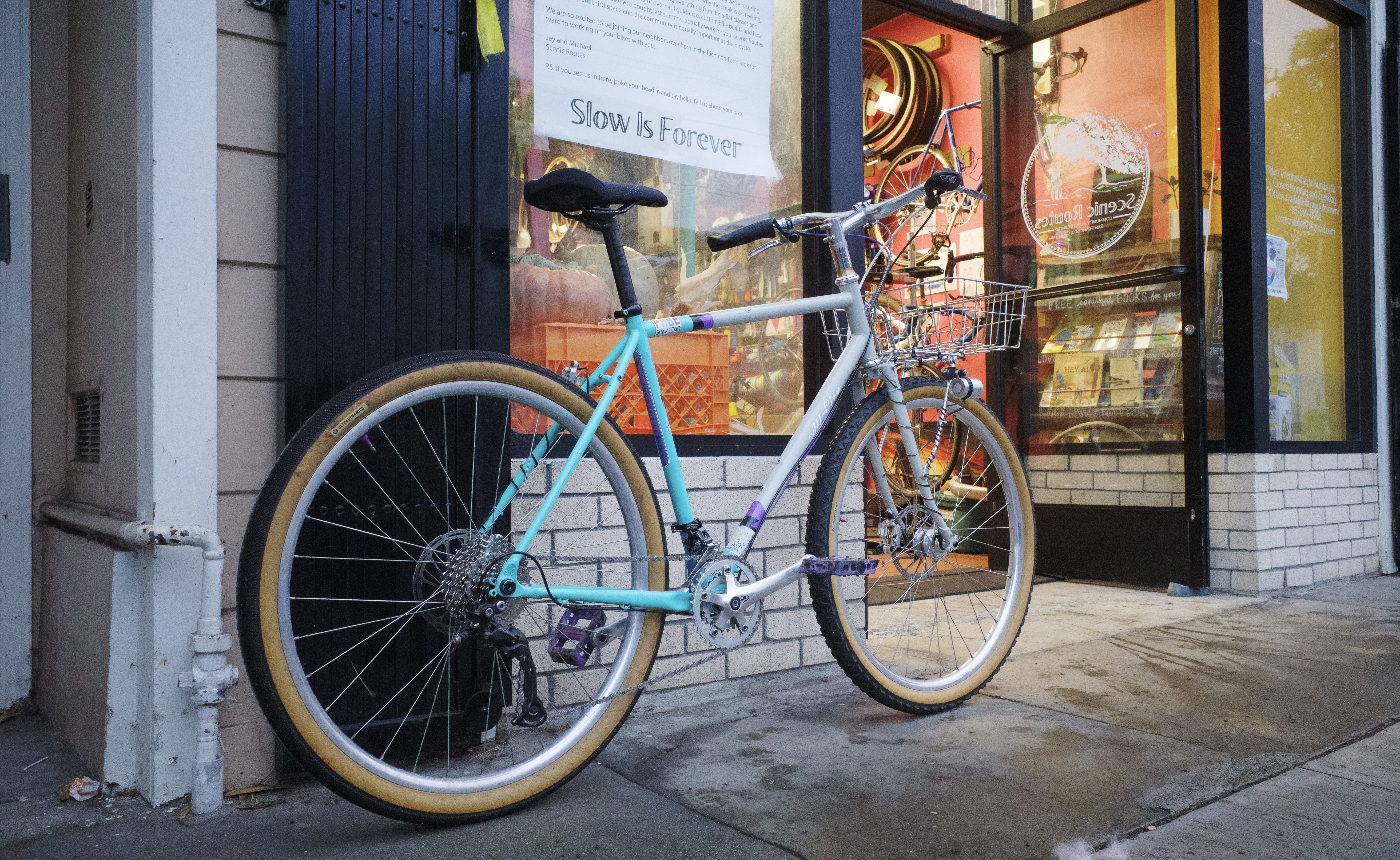 A bicycle with a grey/turquoise frame, black and tan tires, and a mix of black and silver components. It's leaning outside on the wall of Scenic Routes Community Bicycle Center in the evening.