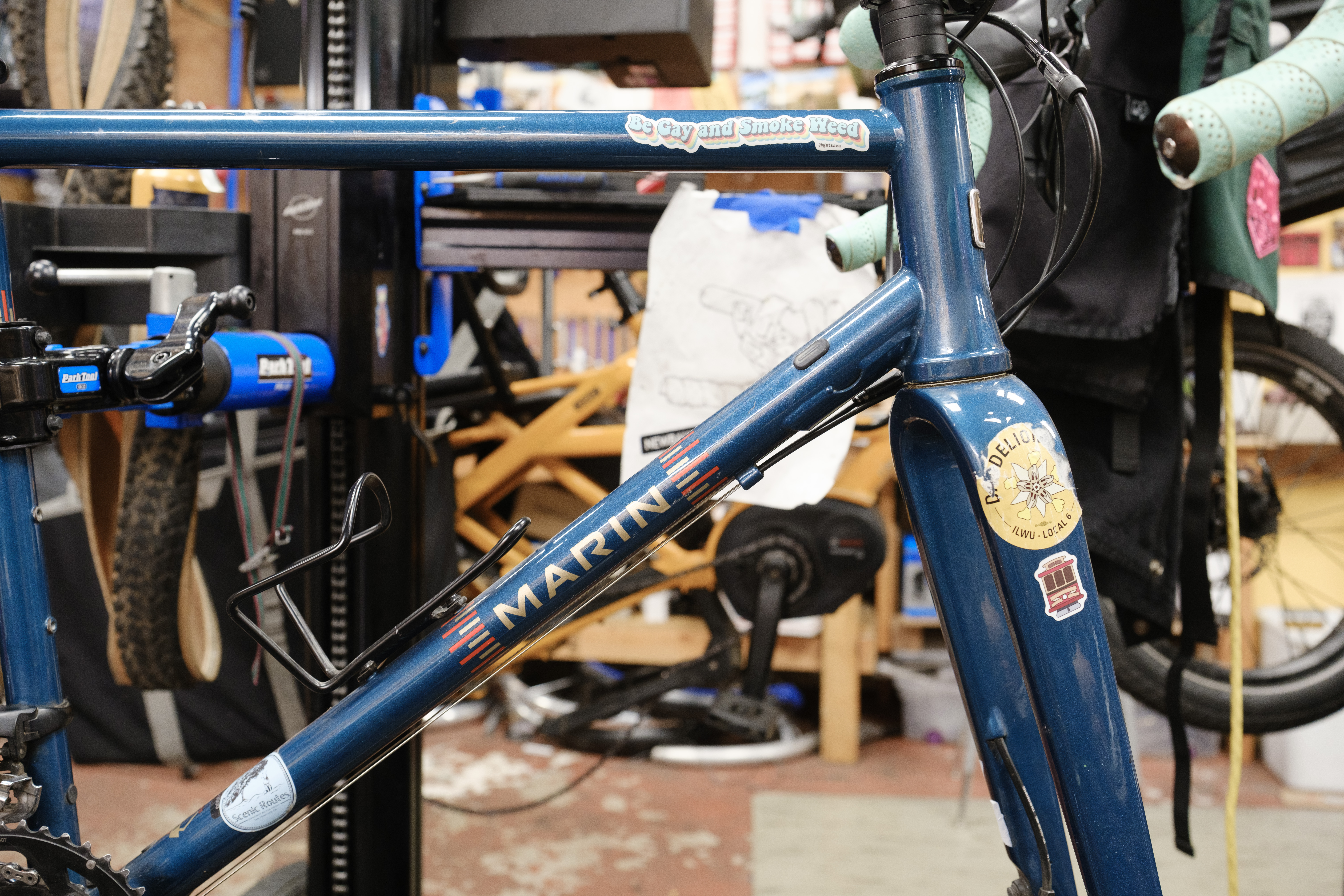 The front triangle of a sparkly blue bicycle in a work stand. The down tube reads 'Marin' and the top tube has a sticker on it that reads 'Be Gay and Smoke Weed'.