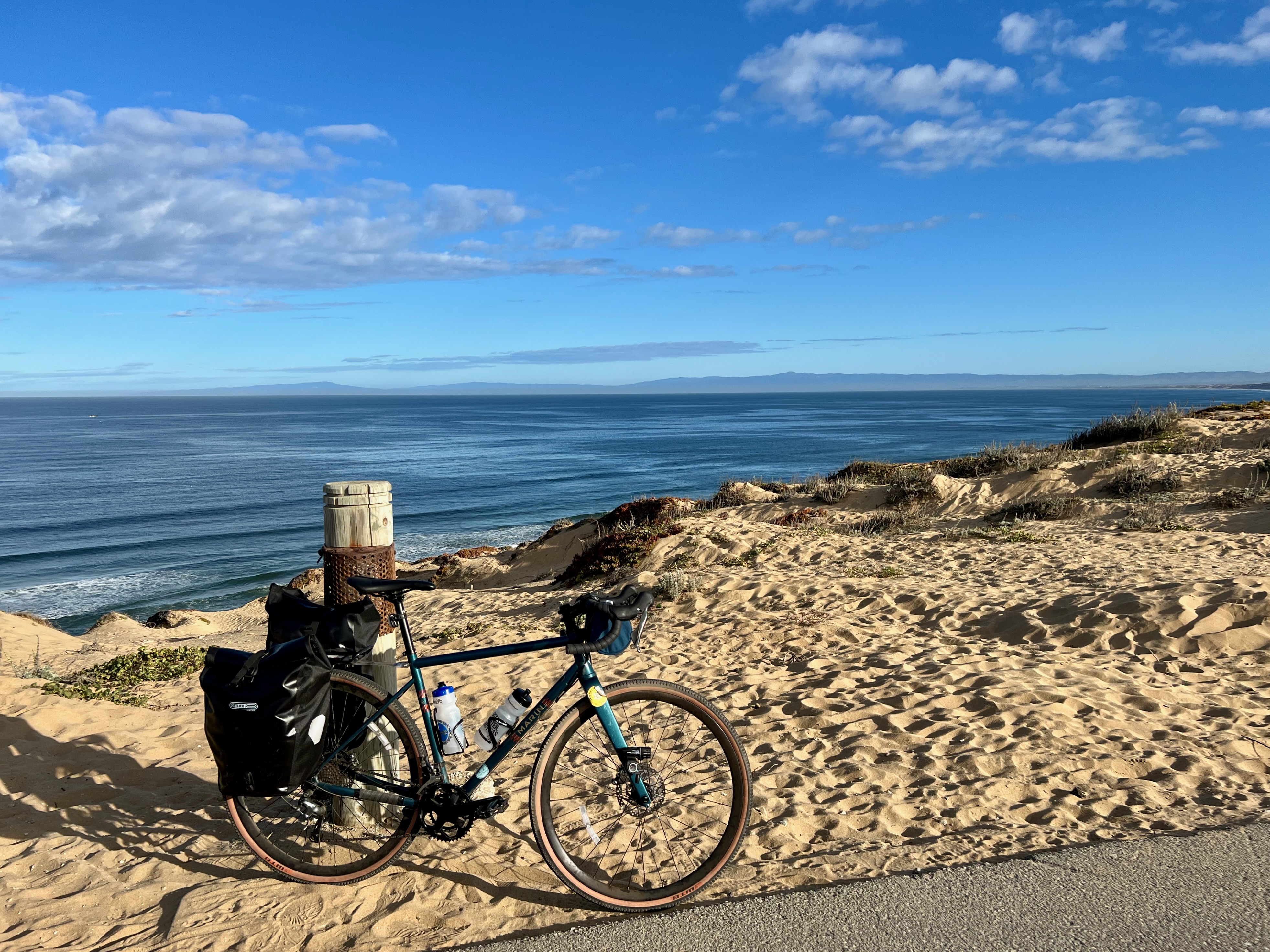 A blue bike with two large panniers on its rear rack leaning against a pole in a sandy dune over the Pacific ocean.