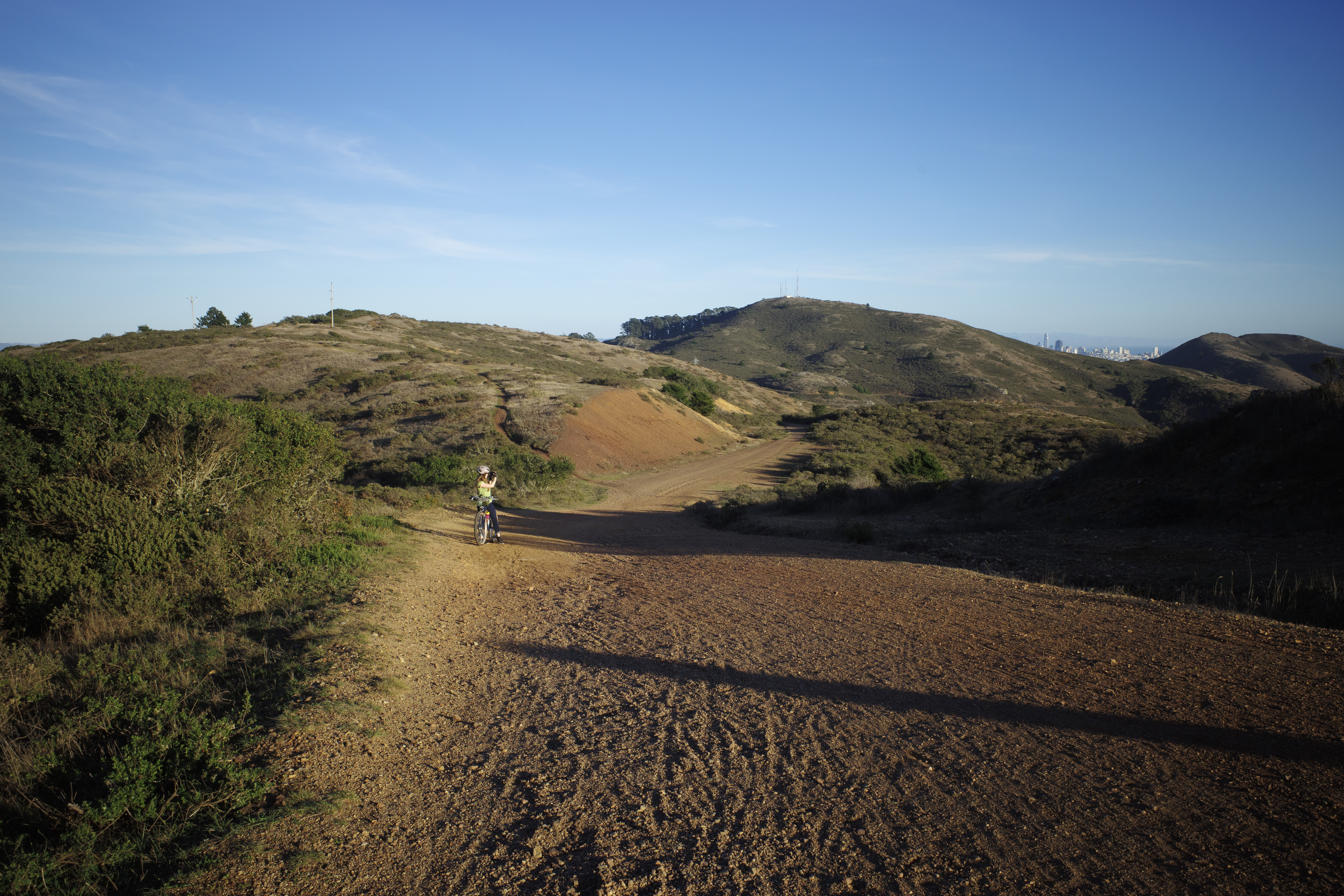 Kat taking a photo toward the ocean on a trail with a bit of the San Francisco skyline in the background