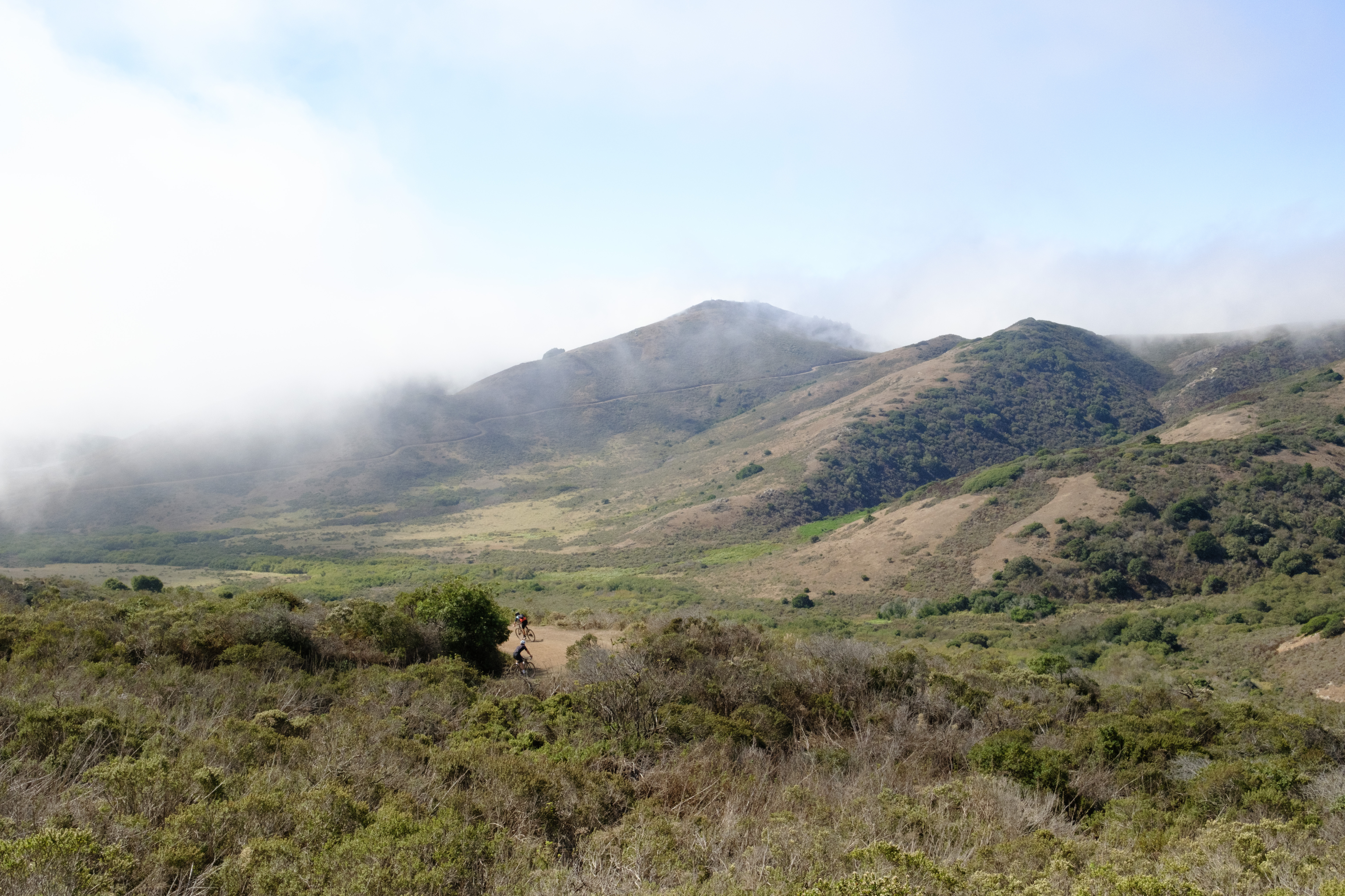 The view down Bobcat with two riders descending around a turn and foggy hills in the background