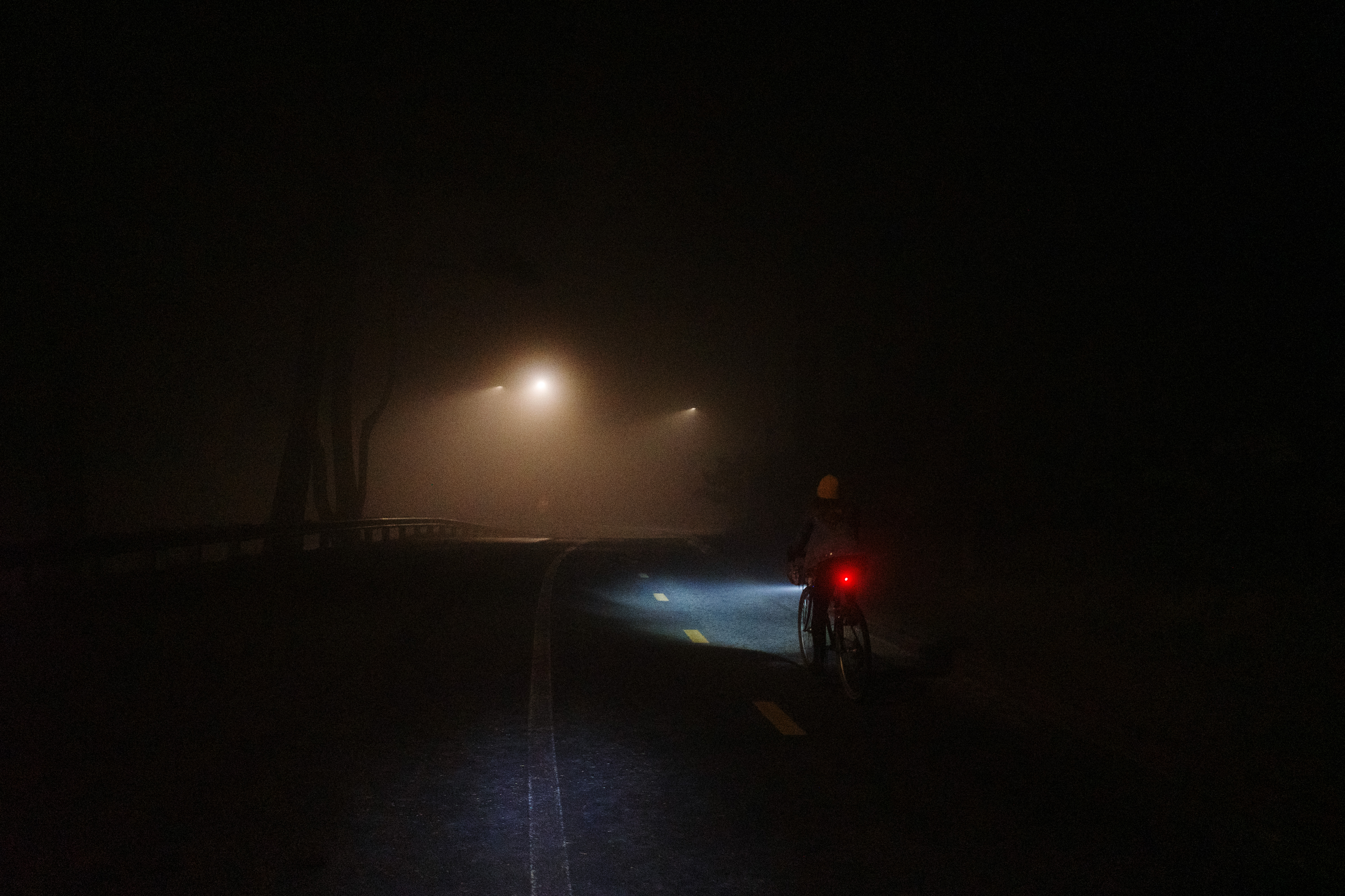 A bicyclist riding down a paved road in the dark with their headlight and a few foggy streetlamps