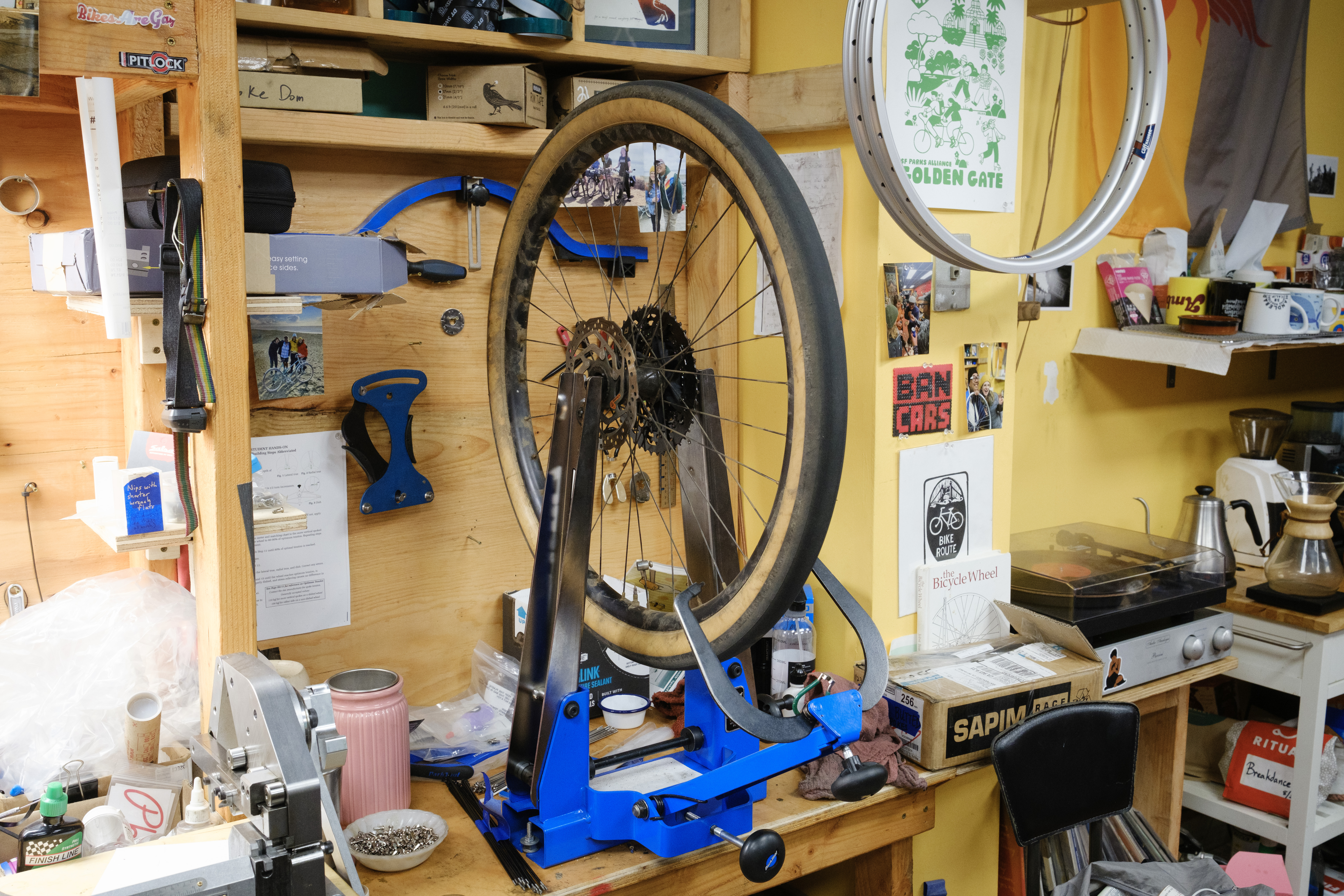 A wheel in a blue truing stand surrounded by a messy bike shop environment.